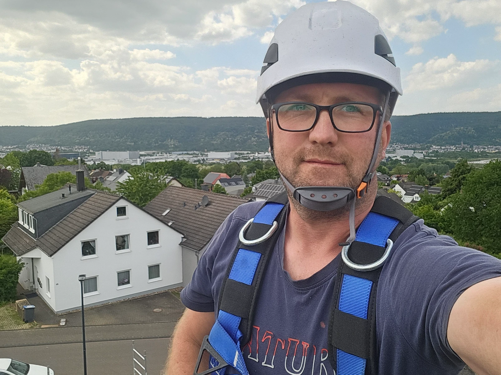 Mann mit Schutzhelm und Gurt auf Dach für Dachreinigung, im Hintergrund Wohnhäuser und grüne Hügel
