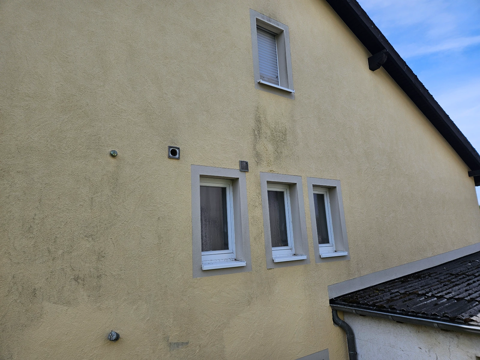 Verunreinigte gelbe Putzfassade mit drei Fenstern und Dachrinne, Fassadenreinigung