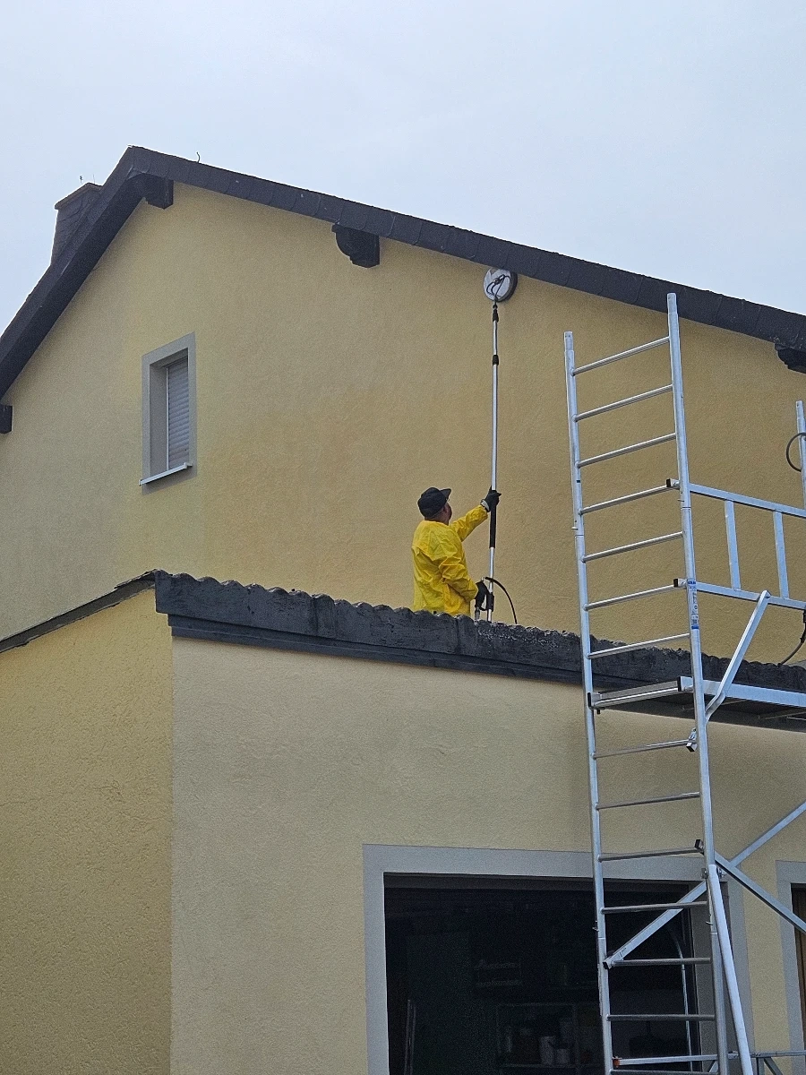 Arbeiter in gelbem Schutzanzug reinigt mit Teleskoplanze die gelbe Hausfassade und Dachüberstand (Fassadenreinigung)