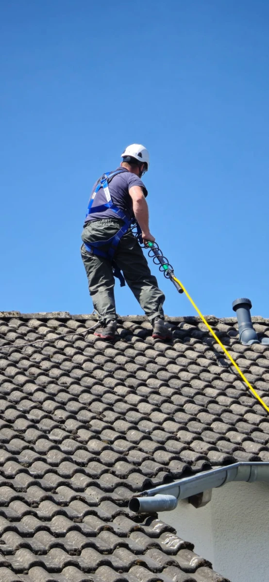 Arbeiter bei Dachreinigung auf Ziegeldach, trägt Schutzhelm und Sicherheitsgurt, bedient Hochdruckschlauch