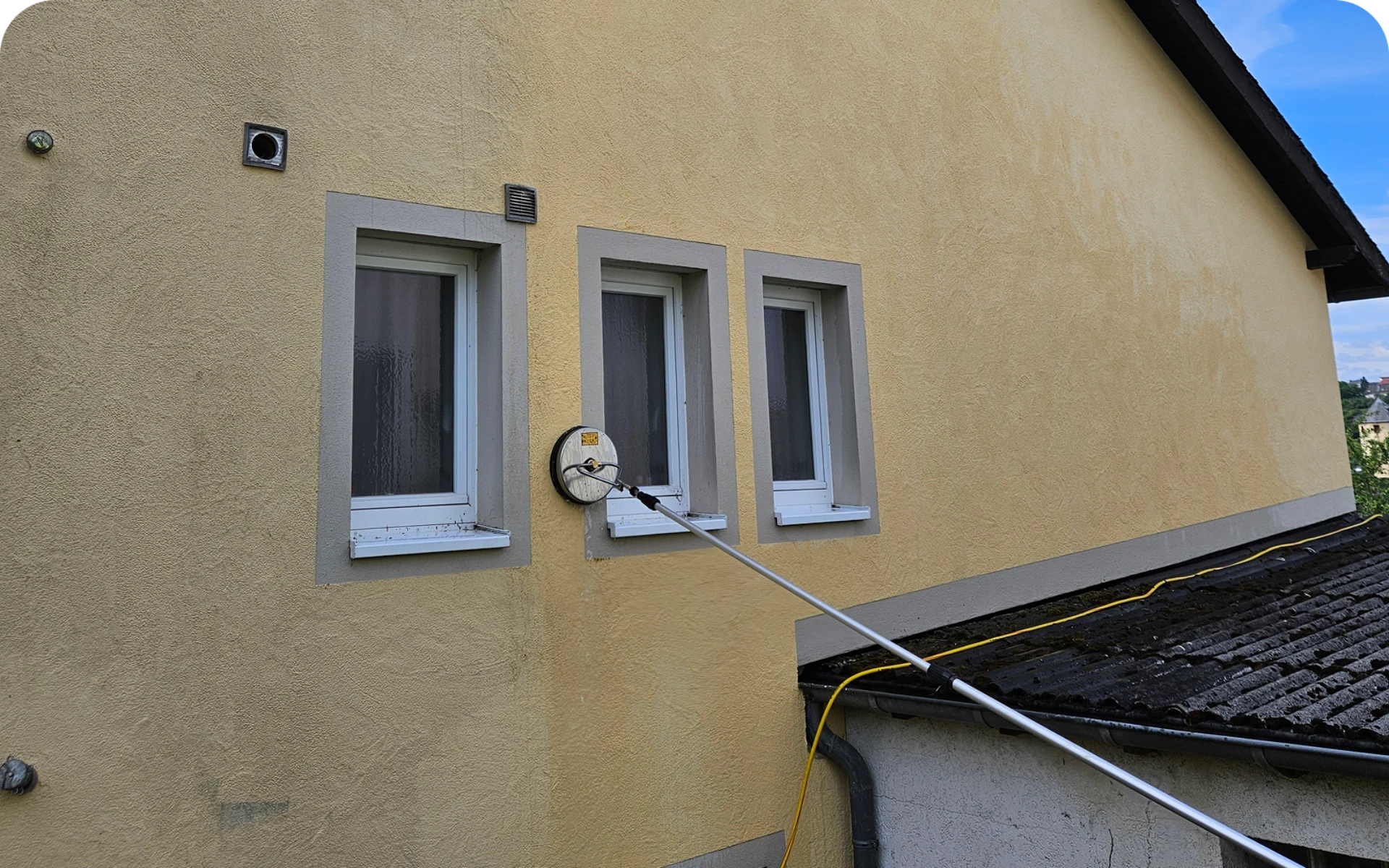 Gelbe Außenfassade mit drei Fenstern und reinigender rotierender Bürstenscheibe an Teleskopstange bei Fassadenreinigung