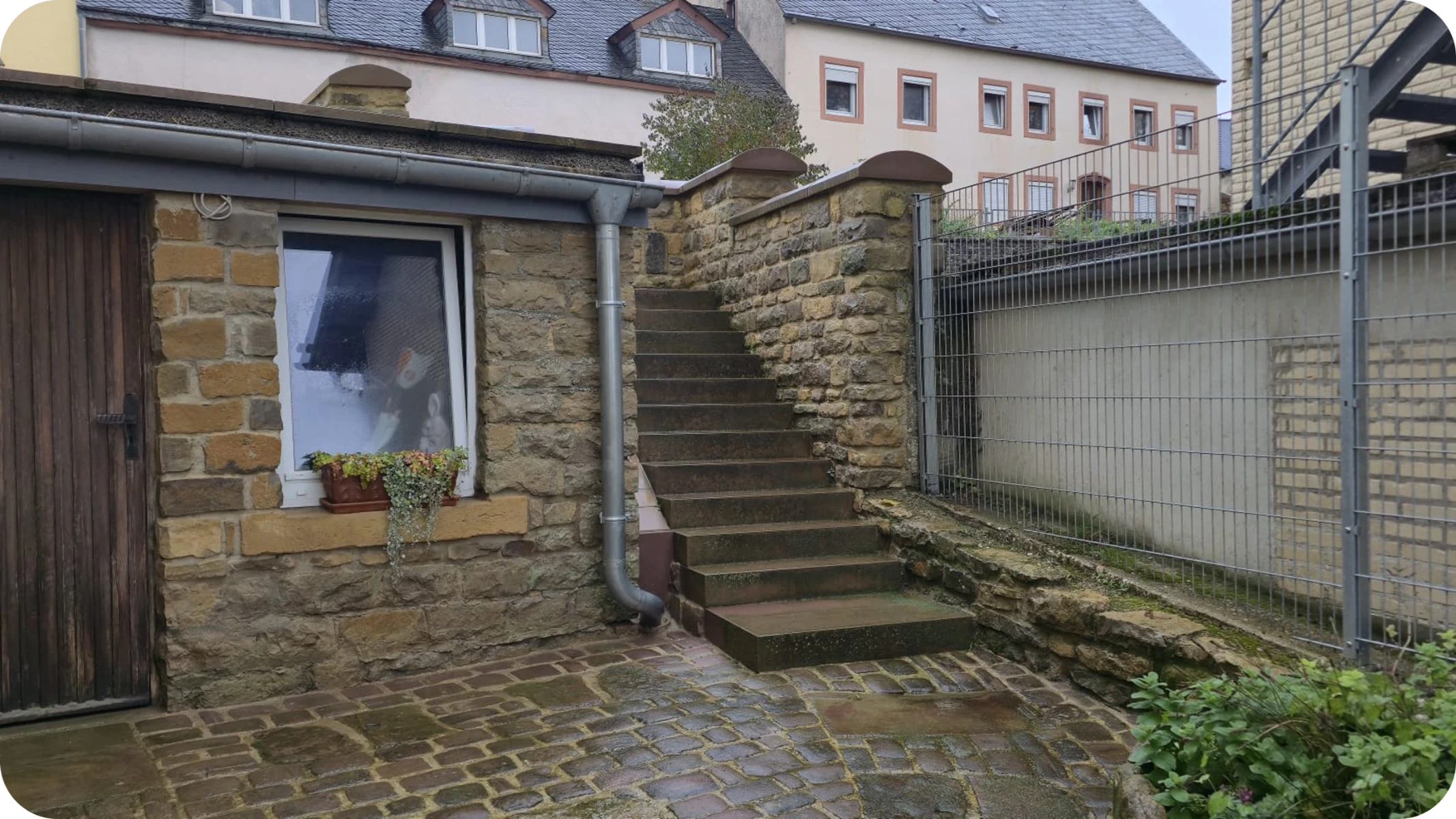 Feuchter Kopfsteinpflasterhof mit Natursteinmauer, Außentreppe und Regenrinne nach Hofreinigung